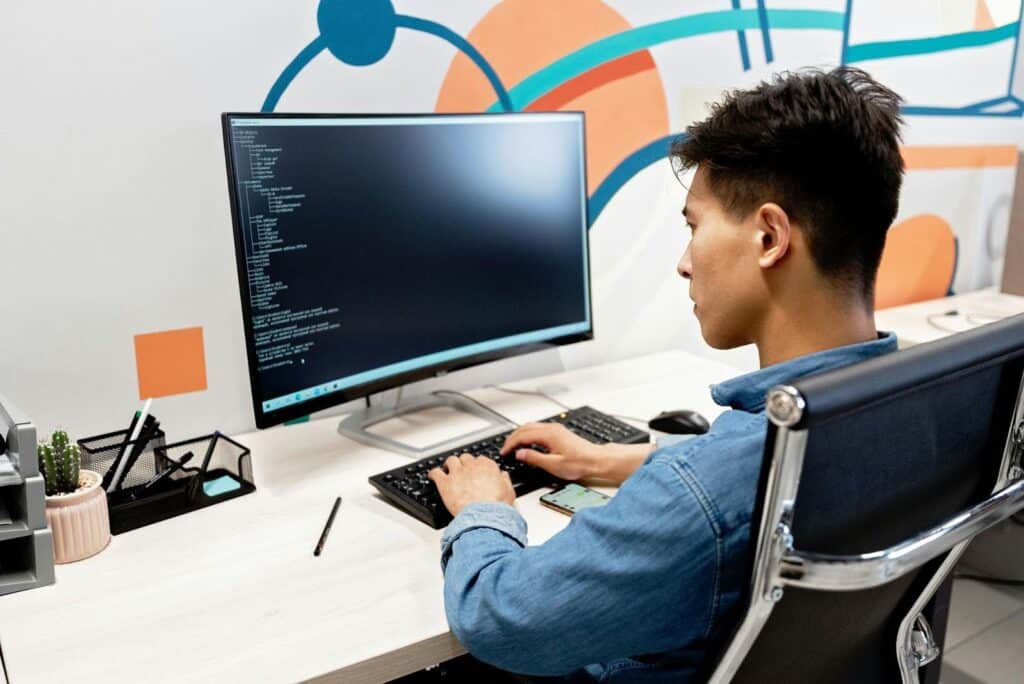 Photo of a man sitting at a desk in an office, typing and looking at a computer screen displaying code.